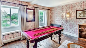 Ein Billardtisch in einem Raum mit ornamentalem Wandpapier, großen Fenstern und Blick ins Grüne.