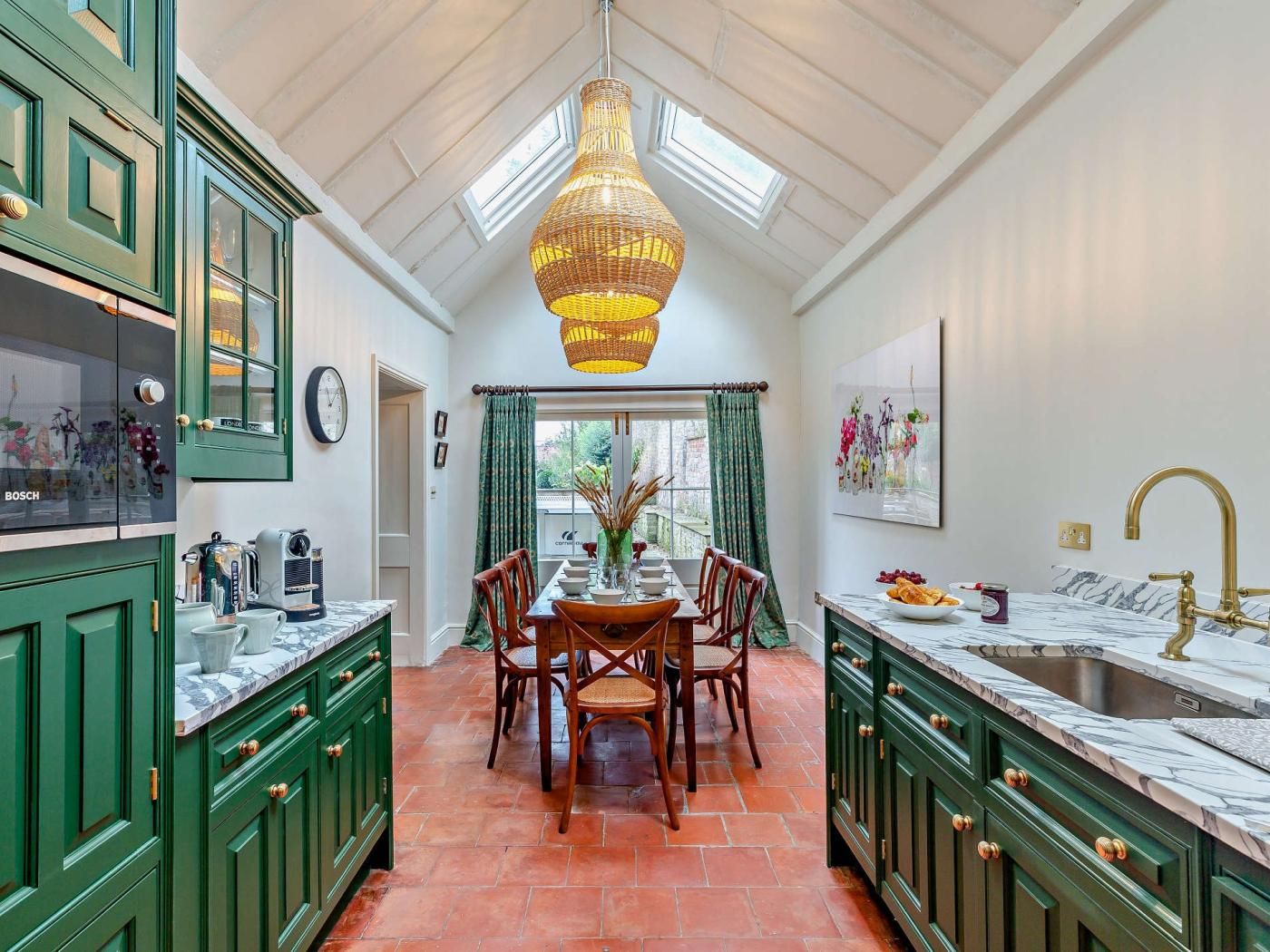 A kitchen with green cabinets, a dining table with chairs, a hanging light fixture, and a window with curtains.