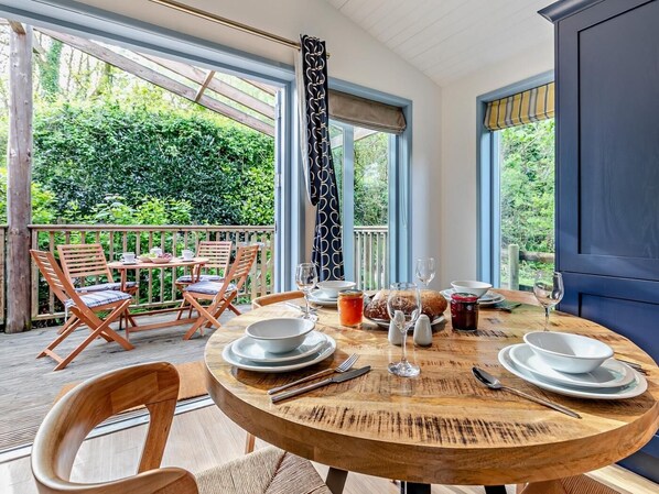 A round wooden table set for dining with chairs, a view of greenery outside, and a large window with curtains.