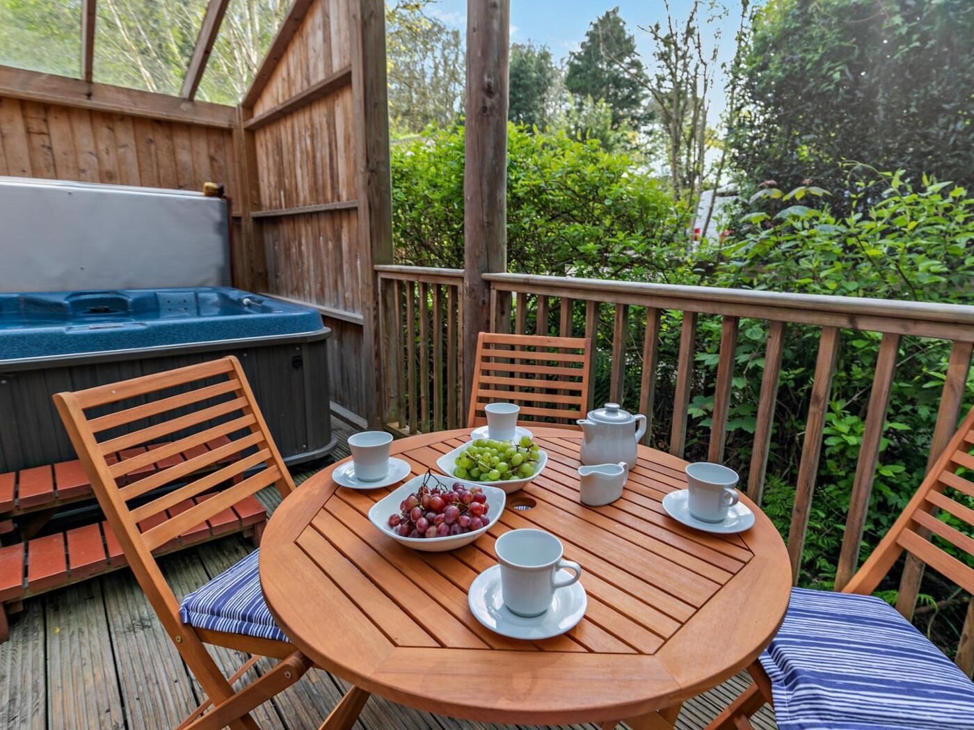 A wooden deck with a hot tub, a round wooden table with chairs, and a view of greenery.