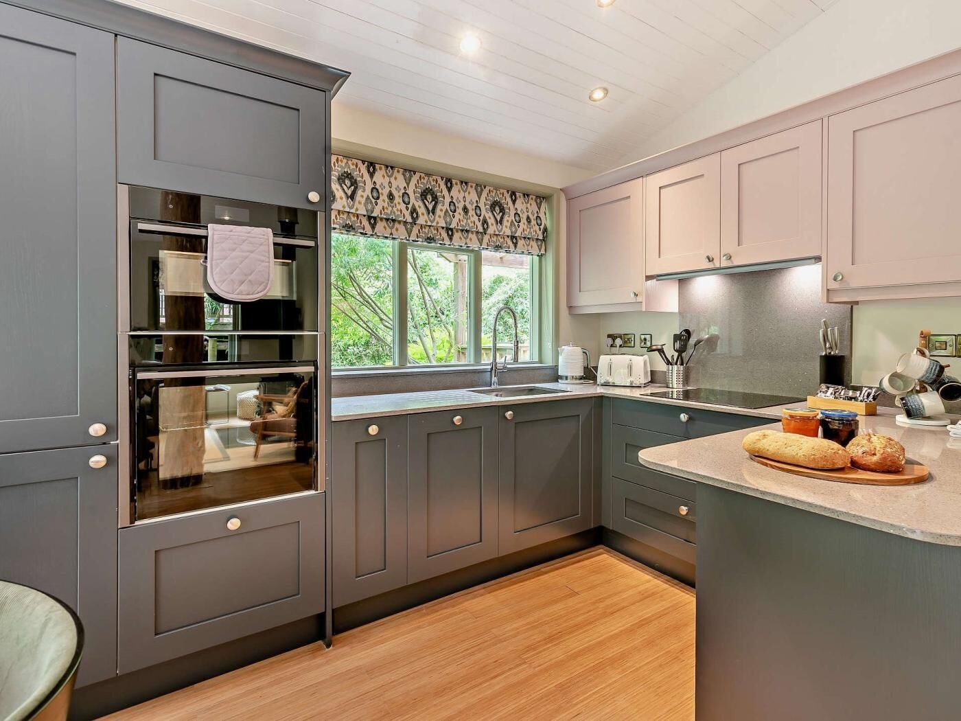 A modern kitchen with grey cabinetry, a built-in oven, a window with a view of greenery, and a countertop with bread and jars.
