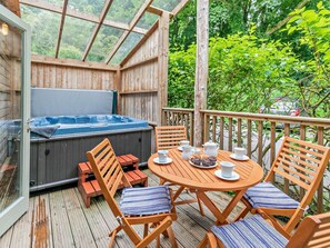 A wooden deck with a hot tub, outdoor seating, and a view of greenery.