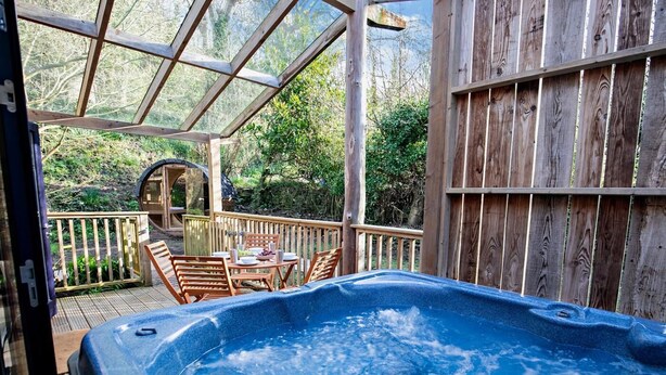 Ein Whirlpool auf einer Holzterrasse mit Glasverkleidung, umgeben von Holzmöbeln und einem Holenzaun.