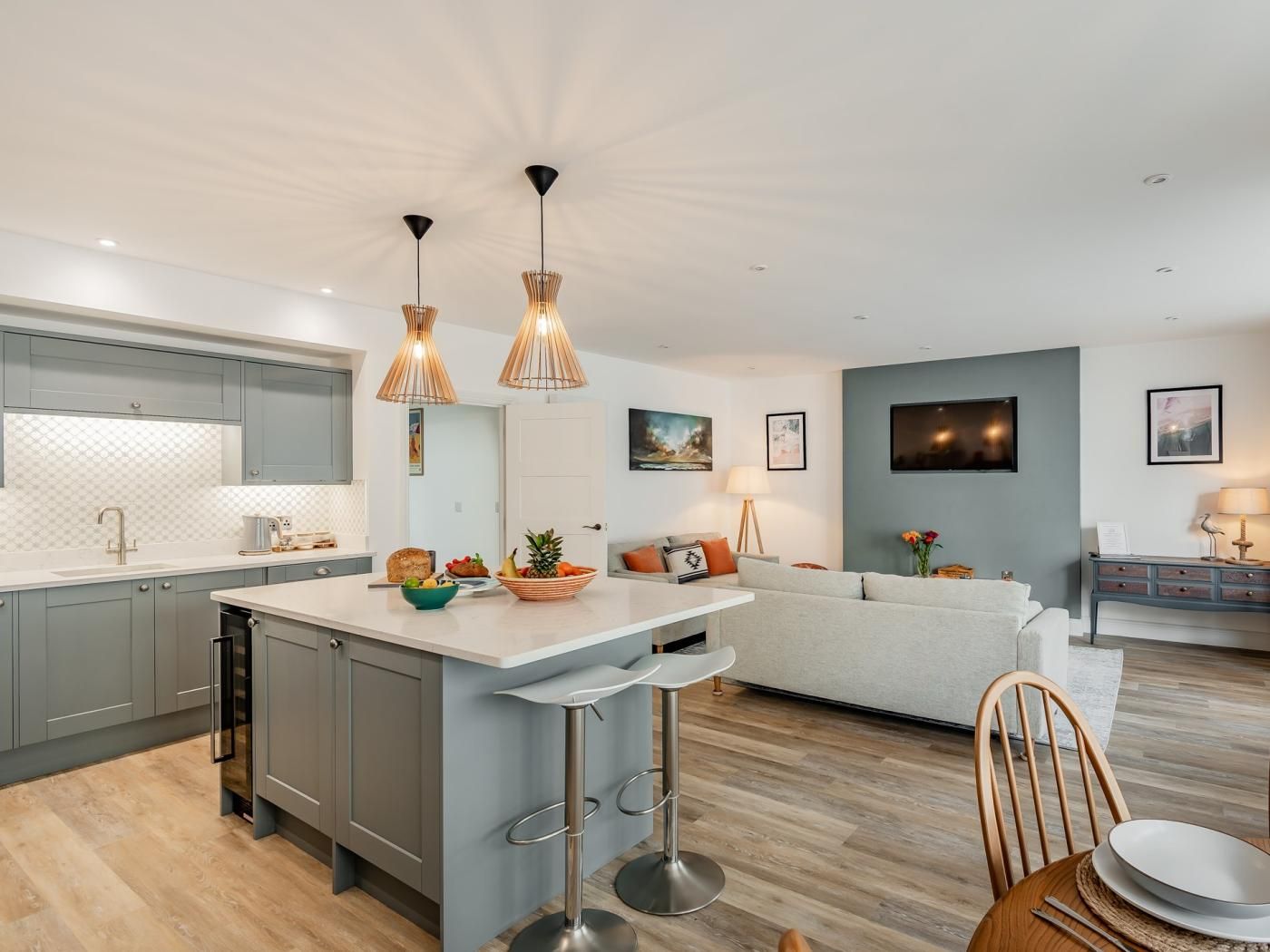 A modern kitchen with grey cabinets, a central island with stools, pendant lighting, and a dining area with a table and chairs.