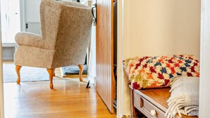 Ein Zimmer mit Holzboden, einem beigefarbenen Sessel, einem hölzernen Bett mit bunter Decke und einem Holzschrank.