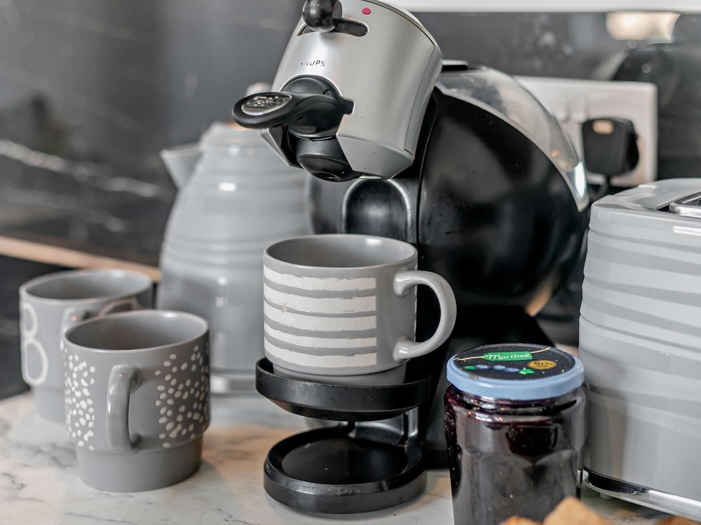 En kaffebryggare, ett glas med sylt och flera muggar står på en köksbänk.
