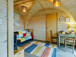 A cozy cabin interior with a wooden ceiling, a bed with colorful bedding, a dining table with chairs, and a small kitchen area.