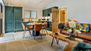 A modern kitchen with teal cabinets, a dining area with a round table and chairs, and a living room with a sofa and a coffee table. - 3 Bed in King's Lynn (oc-93412) (Middleton)