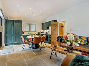 A modern kitchen with teal cabinets, a dining area with a round table and chairs, and a living room with a sofa and a coffee table. - 3 Bed in King's Lynn (oc-93412) (Middleton)