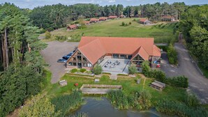 A lodge with a large parking area, a pond, and a forested backdrop. - 3 Bed in King's Lynn (oc-93413) (Middleton)