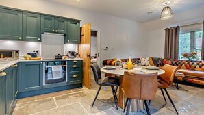 Een moderne keuken met blauwgroene kastjes, een eethoek met een ronde tafel en stoelen, en een gezellige zithoek met een gestreept sofa.
