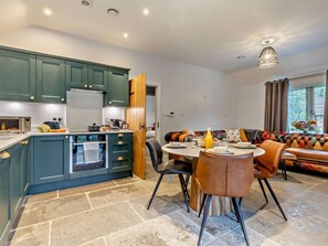 A modern kitchen with teal cabinets, a dining area with a round table and chairs, and a cozy seating area with a patterned sofa. - 3 Bed in King's Lynn (oc-93415) (Middleton)