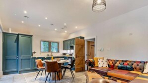 Een ruime woonkamer met een leren bank, een eettafel met stoelen, en een keuken met blauwe kastjes en een houten deur.
