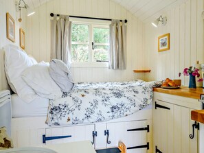 A cozy room with a bed, a window with curtains, a wooden table with a vase of flowers, and a small shelf with a picture.