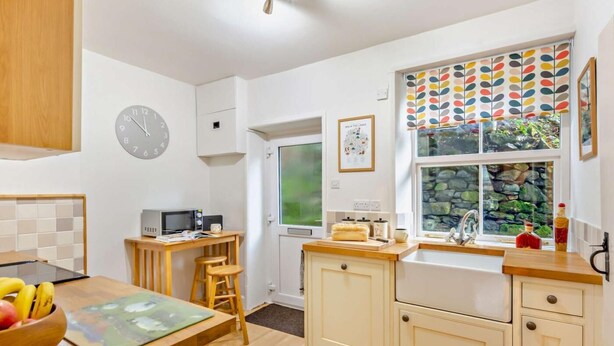 A kitchen with a wooden table, a microwave, a clock, a window with a view, and a sink. - 2 Bed in Penrith (oc-93492) (Glenridding)