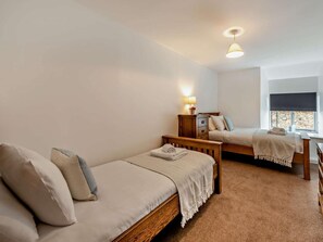 A room with two single beds, a wooden nightstand with a lamp, a window with a blind, and a carpeted floor. - 7 Bed in Bideford (oc-93523) (Weare Giffard)
