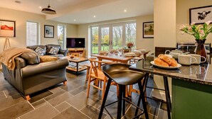 A modern living room with a leather sofa, a dining area with a table and chairs, a kitchen with a counter, and large windows offering a view of the outdoors. - 3 Bed in Keswick (oc-93589) (Threlkeld)