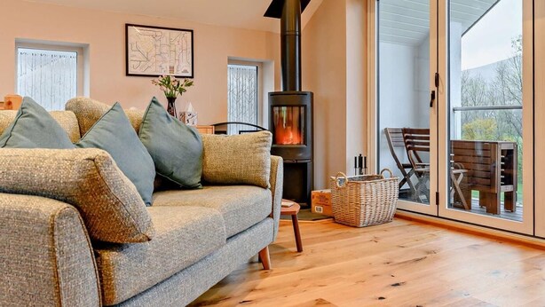 A cozy living room with a sofa, a fireplace, a dining area, and a large window. - 3 Bed in Keswick (oc-93589) (Threlkeld)