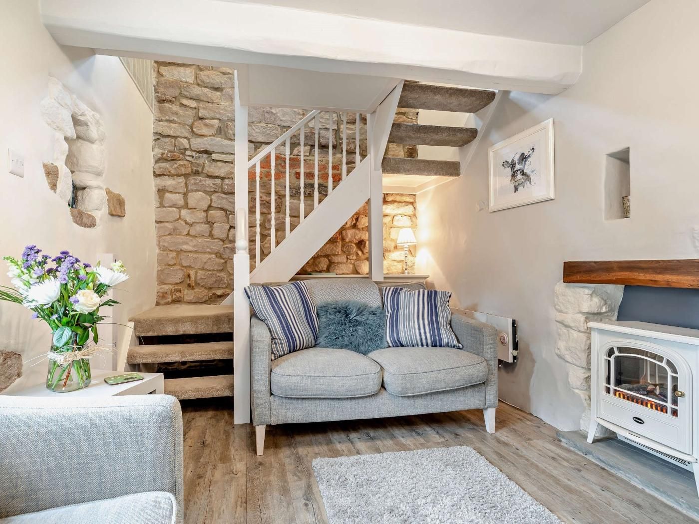 A cozy living room with a stone wall, a staircase, a sofa with striped cushions, and a white fireplace.