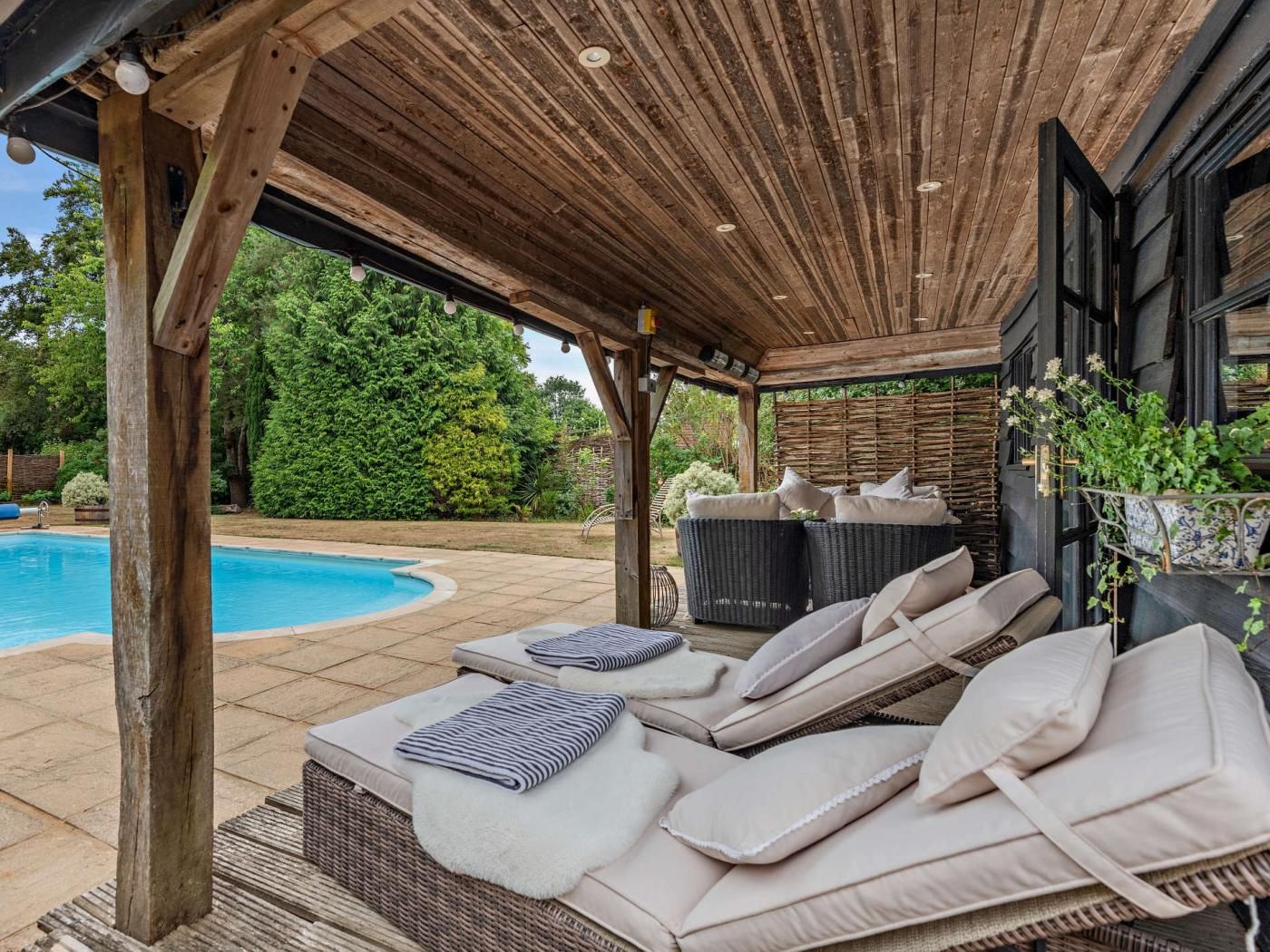 Un espacio de estar al aire libre cubierto con sillones reclinables y cojines, una piscina y una estructura de pérgola de madera.