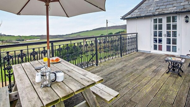 Una terraza de madera con una mesa y un banco, un edificio blanco con techo de tejas y vistas a campos verdes.