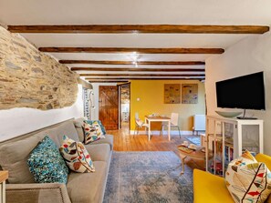 A cozy living room with a stone wall, wooden beams, a sofa with colorful cushions, a television, and a dining area in the background. - 1 Bed in Yelverton (oc-93333) (Yelverton)