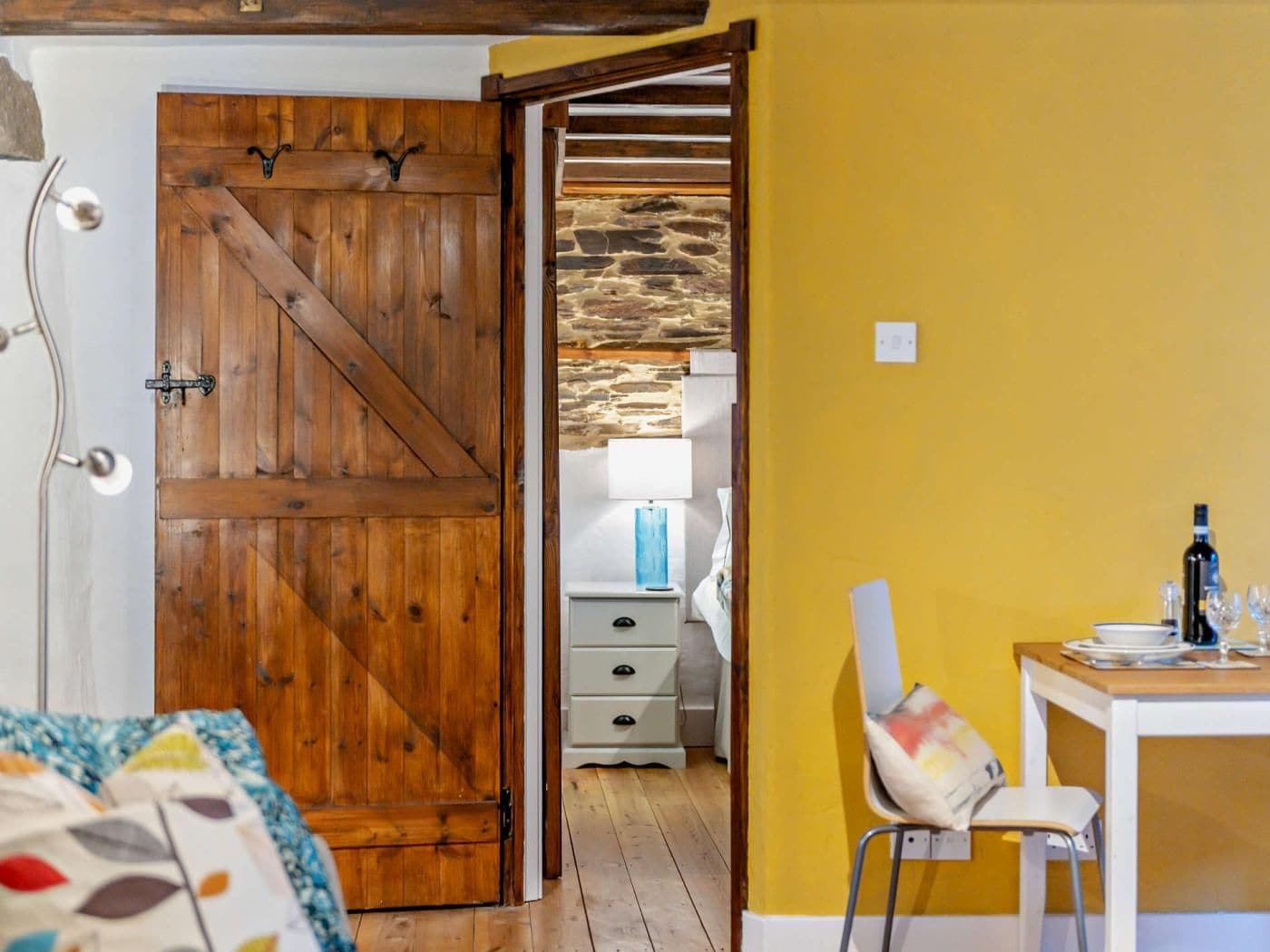 Ein Zimmer mit einer Holztür im Stil eines Bauernhauses, einer Steinwand, einem Bett mit einer bunten Decke, einem kleinen Tisch mit einer Flasche und Gläsern sowie einem Stuhl.