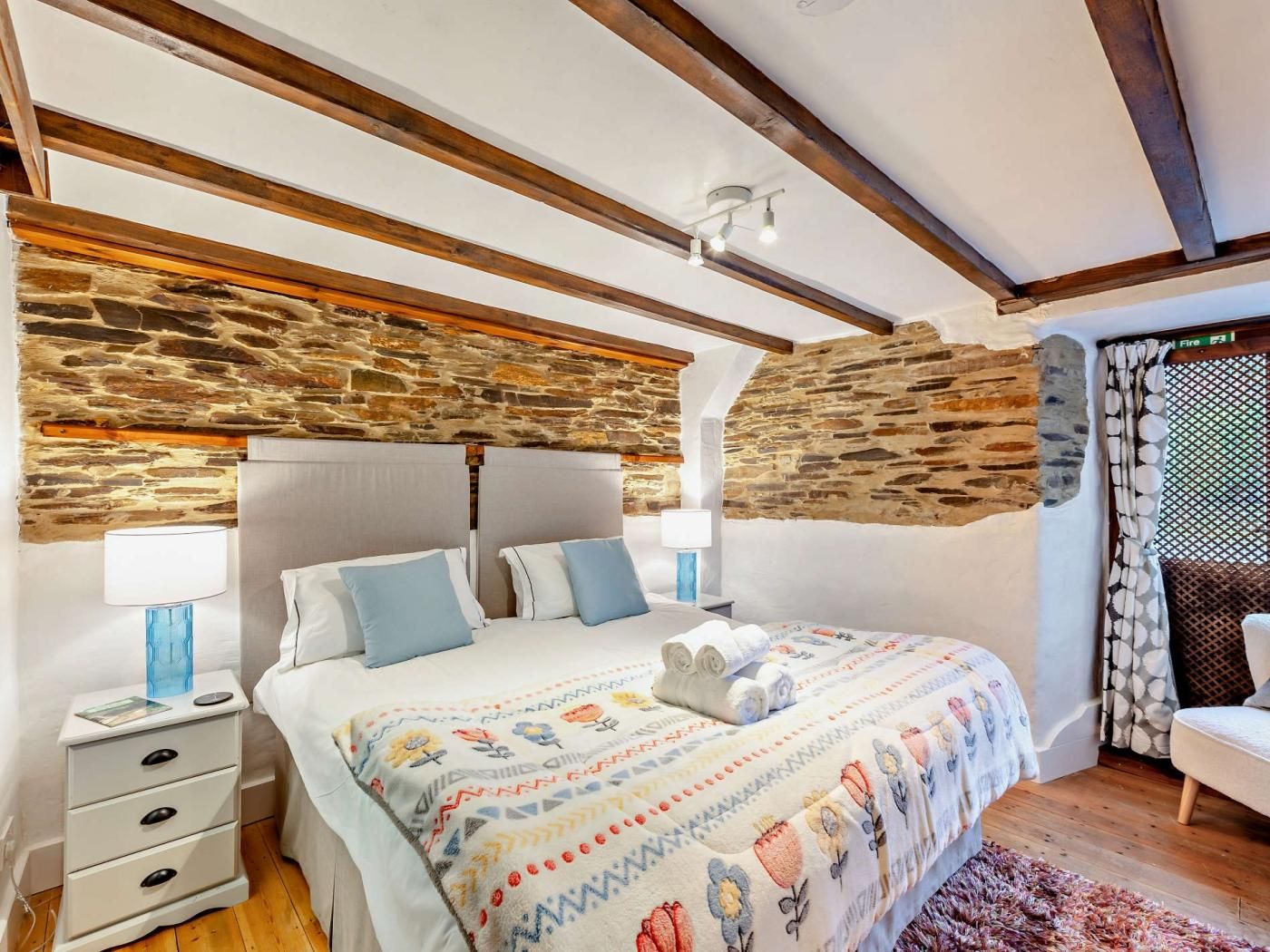 Ein Schlafzimmer mit Steinwand, einem Bett mit gemustertem Bettbezug, Nachttischlampen und einer Holzbalkendecke.