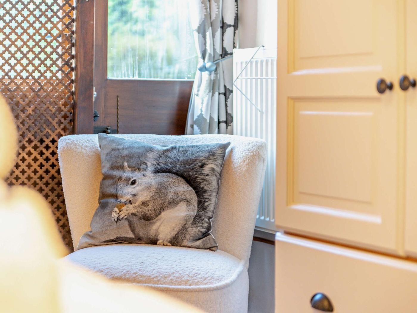 Ein Zimmer mit einem Sessel, einem Kissen mit Eichhörnchen-Motiv, einer Holztür und einem Fenster mit Vorhängen.