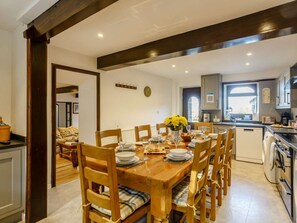 Un comedor con una mesa de madera dispuesta para seis personas, rodeada de sillas con cojines. Se ve un reloj y un gancho en la pared.