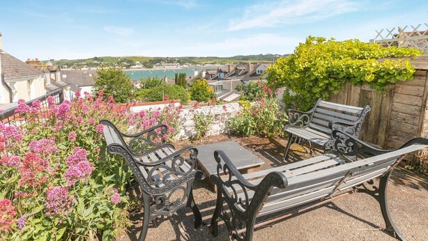 A patio with a table and two benches, surrounded by flowering plants and a view of a coastal town. - 2 Bed in Appledore (oc-gable) (Appledore)