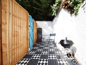 A narrow outdoor patio with a checkered floor, a wooden fence, a blue door, and a barbecue grill.