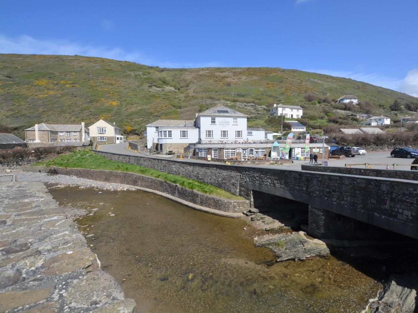 A coastal town with a stone bridge over a shallow water body, leading to a row of white buildings with a sign that reads 'The Coombe Barton.'