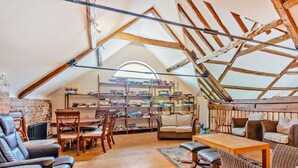 Une pièce mansardée avec un plafond en pente, des poutres en bois, une bibliothèque, des fauteuils en cuir, un canapé et une table à manger.