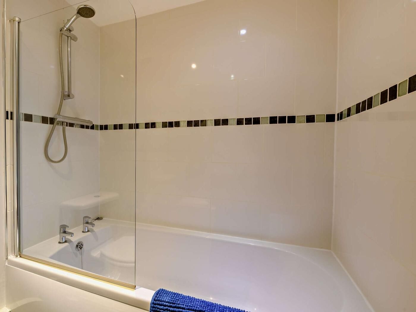 A glass-enclosed shower with a handheld showerhead and a handheld hose, a bathtub, and a blue towel on the floor.