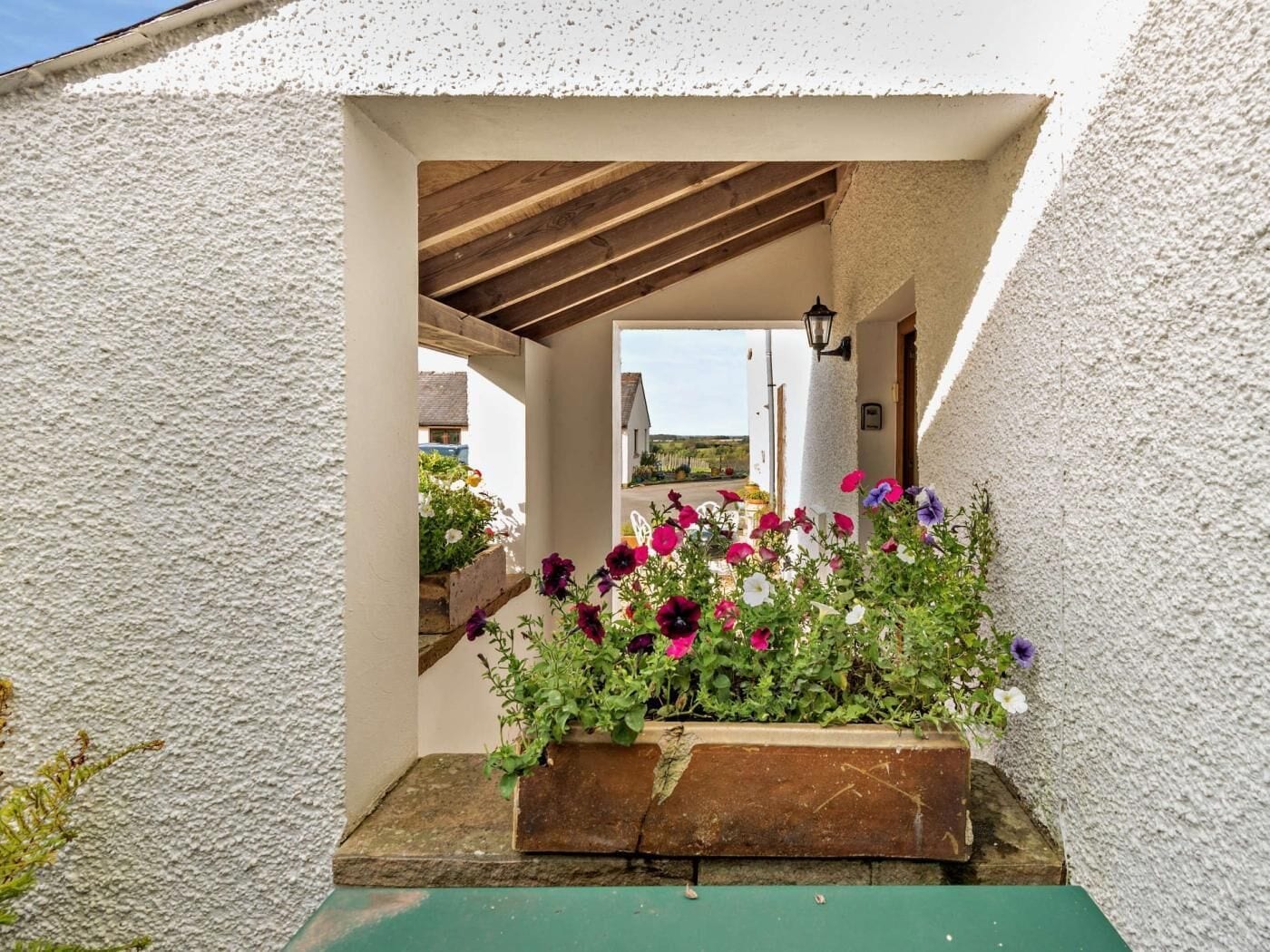 A covered outdoor area with a wooden bench, a planter with flowers, and a view of a building in the background.