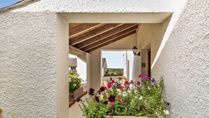 Ein überdachter Außenbereich mit einer Holzbank, einem Blumenbeet und Blick auf ein Gebäude im Hintergrund.