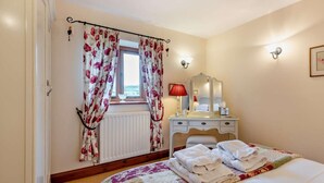 Ein Schlafzimmer mit Bett, Kommode, Spiegel, einem Fenster mit Vorhängen und einer Heizung.