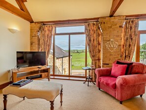 A living room with a television, a red sofa, a wooden bench, and a view of a brick building through large windows. - 2 Bed in Wigton (oc-93463) (Ireby)