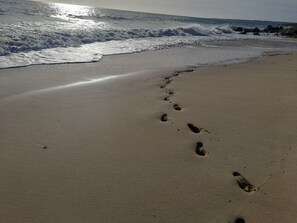 A sandy beach with footprints leading to the water, waves crashing, and a clear sky.