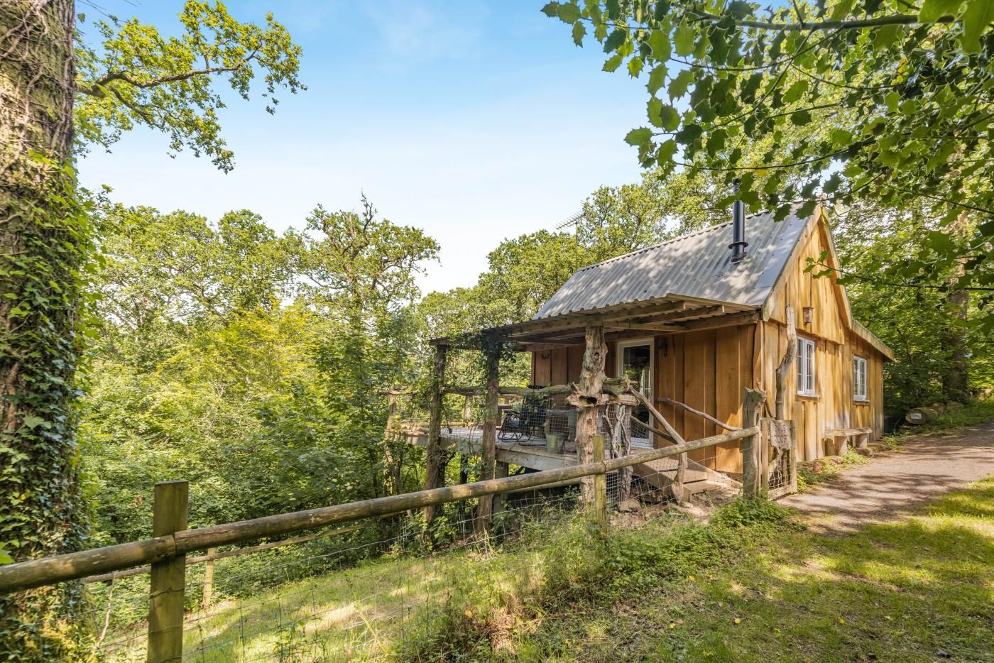 Une cabane en bois rustique, surmontée d’un toit en métal, entourée d’une végétation luxuriante et d’une clôture en bois.