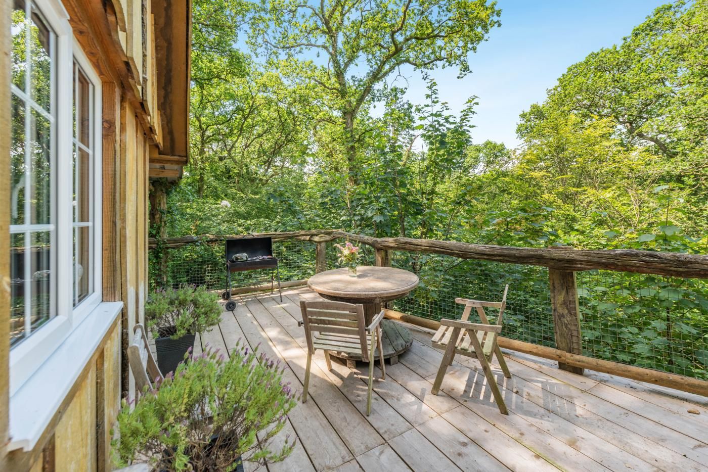 A wooden deck with a round table and chairs, surrounded by a wooden railing, overlooking a dense forest.