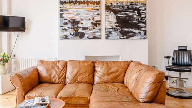 Een bruine leren bank, een rond bijzettafeltje met tijdschriften, en twee abstracte schilderijen aan de muur.