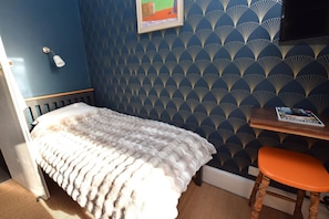 A room with a bed, a small table with a magazine, a chair, and a wall with a geometric pattern. - 7 Bed in St. Leonards-on-sea (oc-52620) (Hastings)