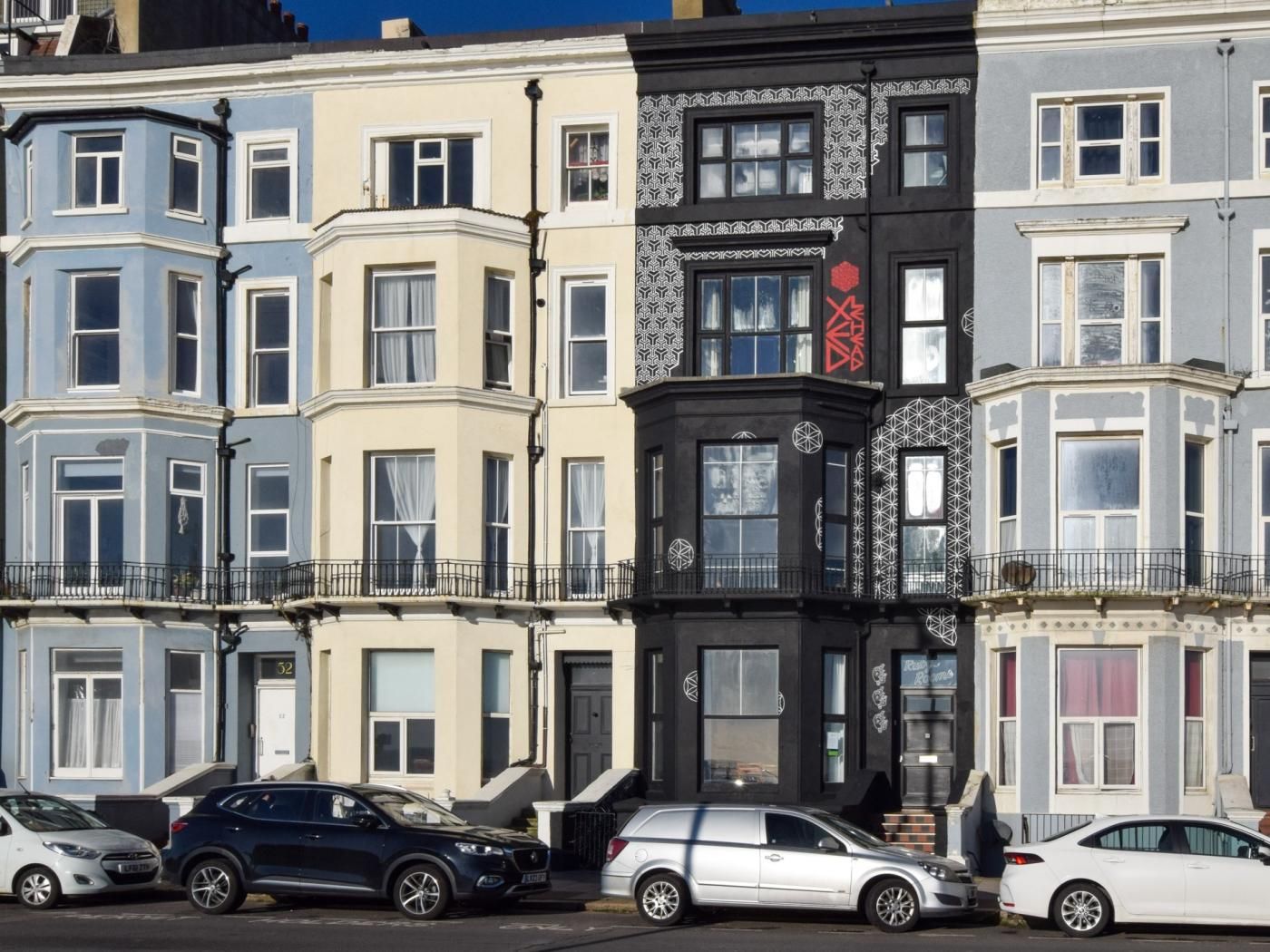 A row of multi-story buildings with bay windows, a black building with red and white patterns, and parked cars.