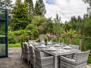 Un espace de restauration en plein air, doté de mobilier en osier et d’une table en verre, entouré d’une végétation luxuriante et d’une clôture en bois.