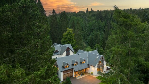 Une grande maison moderne, entourée d’une forêt dense, avec un garage imposant et de nombreuses fenêtres.