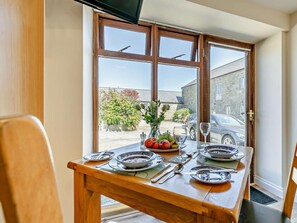 A dining table set with plates, cutlery, and a vase of flowers, positioned in front of a window with a view of a car and buildings. - 1 Bed in Penzance (oc-75689) (Lamorna)