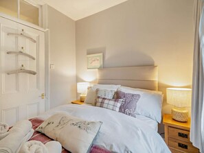 Ein ordentlich bezogenes Bett mit Kissen und Decke, ein Nachttisch mit Lampe sowie eine Tür mit Kleiderhaken.