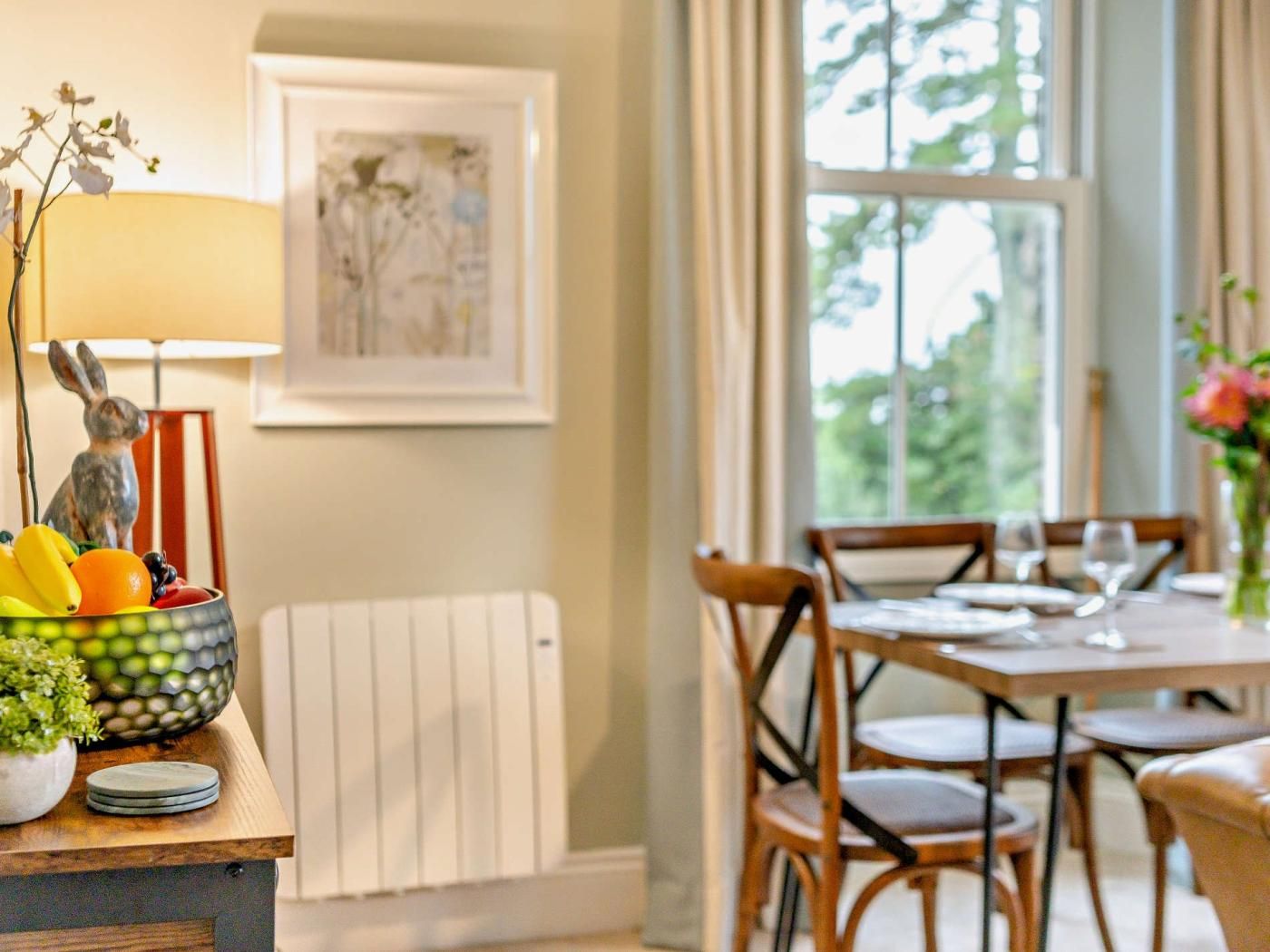 Un comedor con una mesa preparada para cuatro personas, un radiador, una obra de arte enmarcada en la pared y una ventana que da a vistas de árboles.